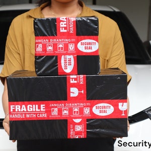 K&ouml;nnte beinhalten: Zwei schwarze Pakete mit rotem Klebeband und wei&szlig;em Text werden vor einem wei&szlig;en Auto gehalten. Die Pakete tragen die Aufschriften "FRAGILE HANDLE WITH CARE" und "SECURITY SEAL". Der Text "JANGAN DIBANTING" ist ebenfalls sichtbar.