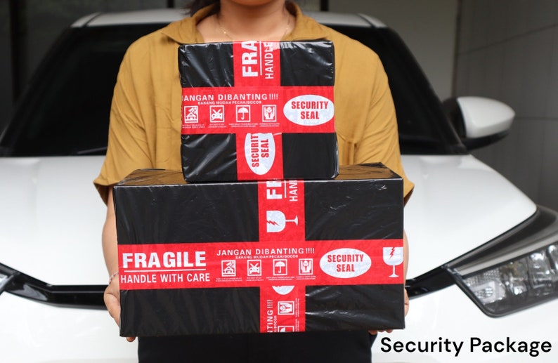 K&ouml;nnte beinhalten: Zwei schwarze Pakete mit rotem Klebeband und wei&szlig;em Text werden vor einem wei&szlig;en Auto gehalten. Die Pakete tragen die Aufschriften "FRAGILE HANDLE WITH CARE" und "SECURITY SEAL". Der Text "JANGAN DIBANTING" ist ebenfalls sichtbar.