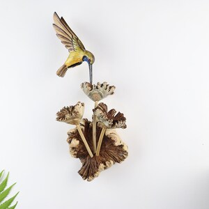 K&ouml;nnte beinhalten: Holzvogel-Skulptur eines Kolibris mit ausgebreiteten Fl&uuml;geln, der sich von einer Blume ern&auml;hrt. Der Vogel hat gelbe, gr&uuml;ne und braune Farbt&ouml;ne, w&auml;hrend die Blume und der Sockel eine nat&uuml;rliche Holzfarbe haben. Die Skulptur ist auf wei&szlig;em Hintergrund montiert.
