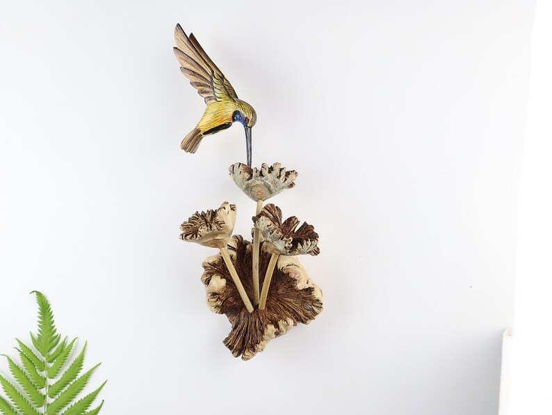 K&ouml;nnte beinhalten: Holzvogel-Skulptur eines Kolibris mit ausgebreiteten Fl&uuml;geln, der sich von einer Blume ern&auml;hrt. Der Vogel hat gelbe, gr&uuml;ne und braune Farbt&ouml;ne, w&auml;hrend die Blume und der Sockel eine nat&uuml;rliche Holzfarbe haben. Die Skulptur ist auf wei&szlig;em Hintergrund montiert.