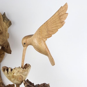 May include: A wooden hummingbird sculpture with outstretched wings, crafted from light-colored wood. The bird is positioned as if drinking from a carved wooden flower. The sculpture is set against a white background, showcasing the natural wood grain.