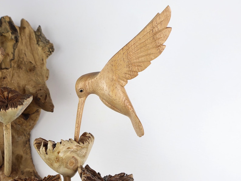 May include: A wooden hummingbird sculpture with outstretched wings, crafted from light-colored wood. The bird is positioned as if drinking from a carved wooden flower. The sculpture is set against a white background, showcasing the natural wood grain.