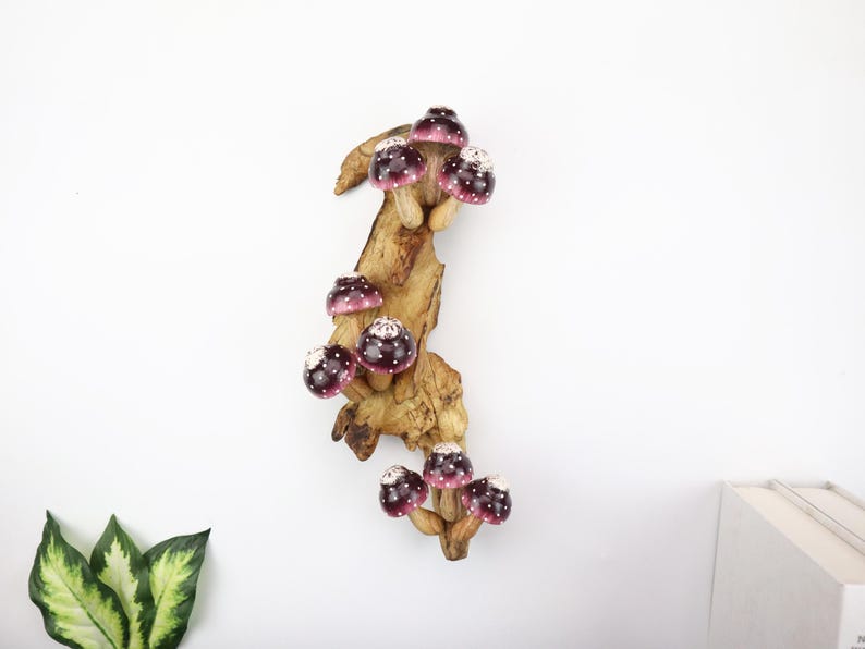 Pu&ograve; includere: Una scultura decorativa in legno con diversi funghi viola e bianchi a pois attaccati a un ramo di legno naturale. I funghi hanno una finitura lucida e sono disposti verticalmente. Il legno ha un aspetto naturale e strutturato.