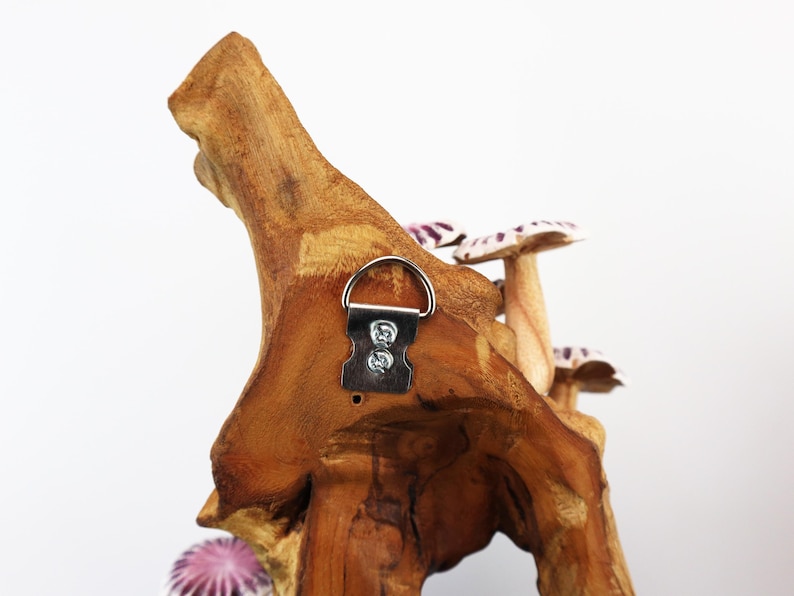 May include: A wooden wall hanging featuring a natural wood base with several mushroom sculptures. The mushrooms have white and purple caps. A metal hanger is attached to the back for easy display.