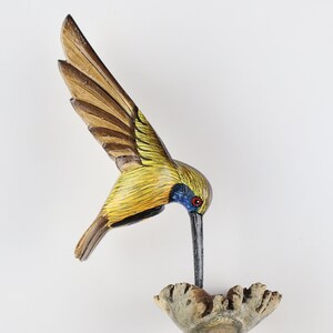 K&ouml;nnte beinhalten: Handgeschnitzte Holzskulptur eines Kolibris mit ausgebreiteten Fl&uuml;geln. Der Vogel weist gelbe, braune und blaue Farbakzente auf. Er sitzt auf einem Holzsockel und scheint daraus zu trinken. Die Skulptur ist ein dekoratives Objekt.