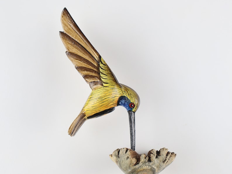 K&ouml;nnte beinhalten: Handgeschnitzte Holzskulptur eines Kolibris mit ausgebreiteten Fl&uuml;geln. Der Vogel weist gelbe, braune und blaue Farbakzente auf. Er sitzt auf einem Holzsockel und scheint daraus zu trinken. Die Skulptur ist ein dekoratives Objekt.