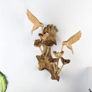 May include: Wooden wall art featuring two carved hummingbirds hovering near stylized flowers. The birds and flowers are light brown, mounted on a textured, dark brown wooden base. A nature-themed decorative piece.