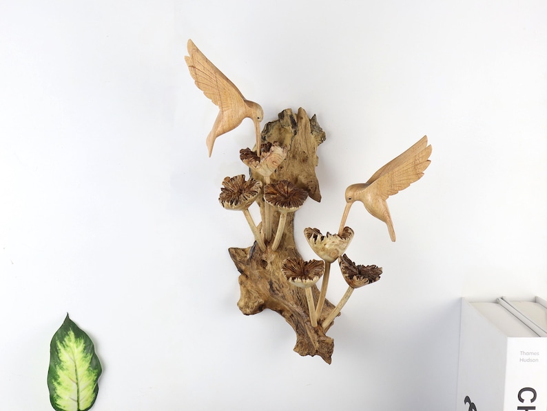 May include: Wooden wall art featuring two carved hummingbirds hovering near stylized flowers. The birds and flowers are light brown, mounted on a textured, dark brown wooden base. A nature-themed decorative piece.