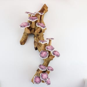May include: A decorative wooden wall hanging featuring a cluster of stylized mushrooms. The mushrooms have pink and white caps and are attached to a natural-toned wooden branch. The artwork is mounted on a white wall.
