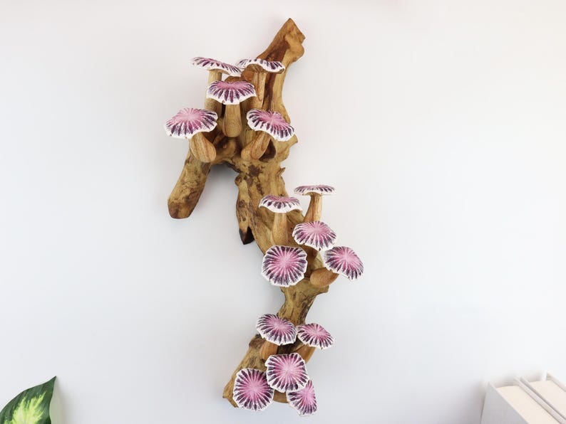May include: A decorative wooden wall hanging featuring a cluster of stylized mushrooms. The mushrooms have pink and white caps and are attached to a natural-toned wooden branch. The artwork is mounted on a white wall.