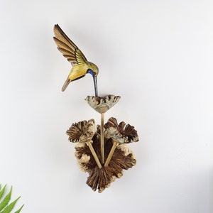 K&ouml;nnte beinhalten: Eine Holzskulptur eines Kolibris mit ausgebreiteten Fl&uuml;geln, bemalt in Braun-, Gelb- und Blaut&ouml;nen, sitzt &uuml;ber einer dekorativen Holzblume. Die Blume besteht aus strukturiertem Holz und hat drei Stiele. Die Skulptur ist an einer wei&szlig;en Wand befestigt.