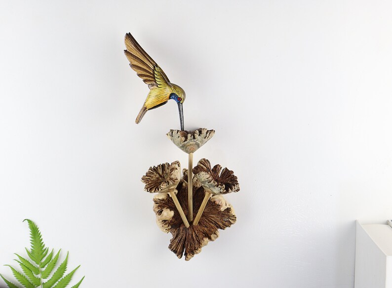 K&ouml;nnte beinhalten: Eine Holzskulptur eines Kolibris mit ausgebreiteten Fl&uuml;geln, bemalt in Braun-, Gelb- und Blaut&ouml;nen, sitzt &uuml;ber einer dekorativen Holzblume. Die Blume besteht aus strukturiertem Holz und hat drei Stiele. Die Skulptur ist an einer wei&szlig;en Wand befestigt.