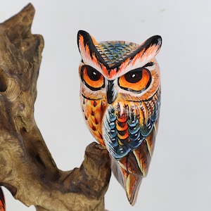 Puede incluir: Una figurita de b&uacute;ho de madera pintada a mano con plumas naranja, azul y marr&oacute;n. El b&uacute;ho est&aacute; posado en una rama con una textura de veta de madera detallada.