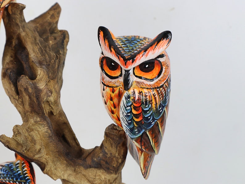 Puede incluir: Una figurita de b&uacute;ho de madera pintada a mano con plumas naranja, azul y marr&oacute;n. El b&uacute;ho est&aacute; posado en una rama con una textura de veta de madera detallada.