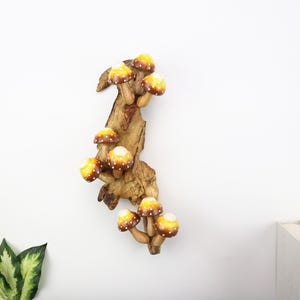 Pu&ograve; includere: Decorazione murale in legno con un gruppo di funghi stilizzati. I funghi hanno cappelli gialli e marroni con pois bianchi, attaccati a un pezzo di legno naturale. Il pezzo &egrave; montato su una parete bianca.