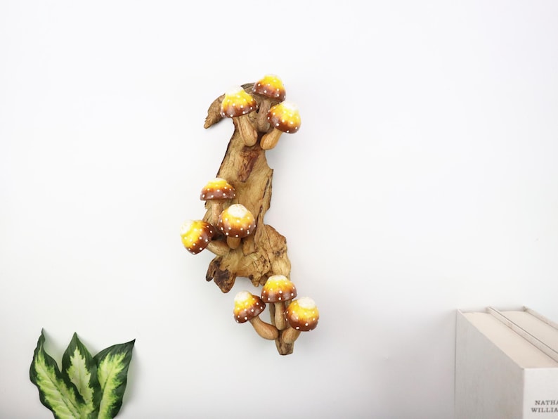 Pu&ograve; includere: Decorazione murale in legno con un gruppo di funghi stilizzati. I funghi hanno cappelli gialli e marroni con pois bianchi, attaccati a un pezzo di legno naturale. Il pezzo &egrave; montato su una parete bianca.