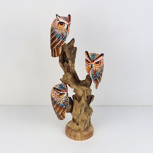 Puede incluir: Tres b&uacute;hos de madera pintados a mano posados ​​en una rama de &aacute;rbol. Los b&uacute;hos est&aacute;n pintados en tonos de naranja, marr&oacute;n, azul y negro. La rama del &aacute;rbol est&aacute; hecha de madera flotante y tiene un color marr&oacute;n natural.