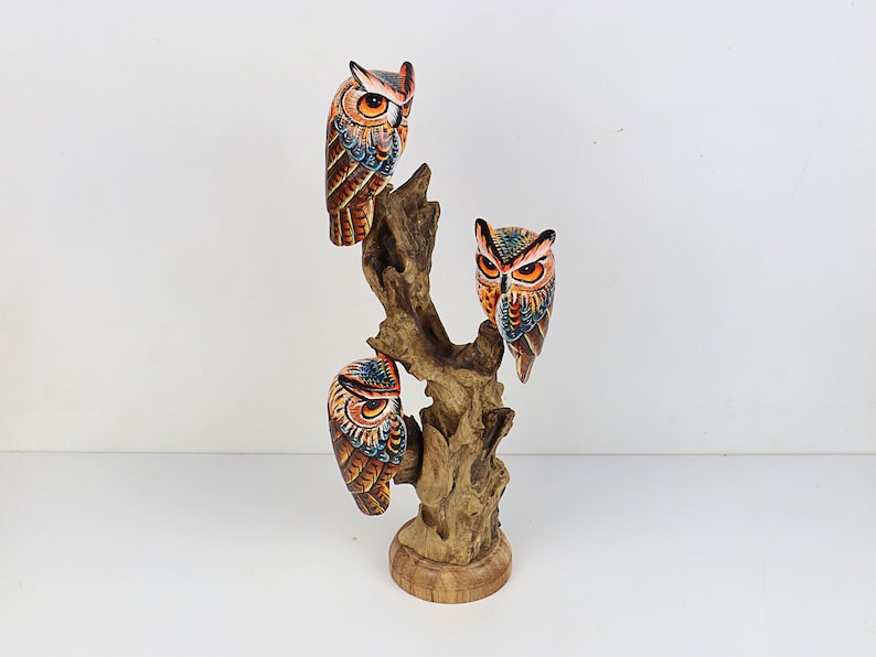 Puede incluir: Tres b&uacute;hos de madera pintados a mano posados ​​en una rama de &aacute;rbol. Los b&uacute;hos est&aacute;n pintados en tonos de naranja, marr&oacute;n, azul y negro. La rama del &aacute;rbol est&aacute; hecha de madera flotante y tiene un color marr&oacute;n natural.