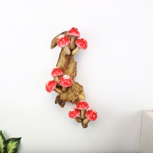 Pu&ograve; includere: Una decorazione da parete in legno con un gruppo di funghi rossi a pois bianchi. I funghi sono attaccati a un pezzo di legno naturale, creando un'estetica stravagante e rustica. Lo sfondo &egrave; bianco.