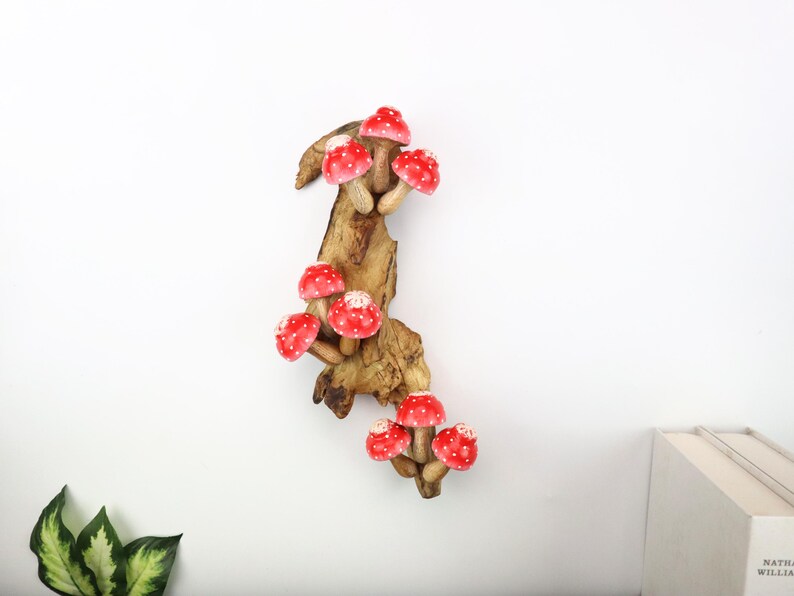 Pu&ograve; includere: Una decorazione da parete in legno con un gruppo di funghi rossi a pois bianchi. I funghi sono attaccati a un pezzo di legno naturale, creando un'estetica stravagante e rustica. Lo sfondo &egrave; bianco.