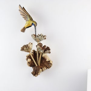 K&ouml;nnte beinhalten: Eine Holzskulptur eines Kolibris in Gelb, Blau und Braun, der &uuml;ber einem Strau&szlig; geschnitzter Holzblumen schwebt. Die Skulptur ist auf einem hellen Holzsockel montiert und schafft ein von der Natur inspiriertes Dekorationsobjekt.
