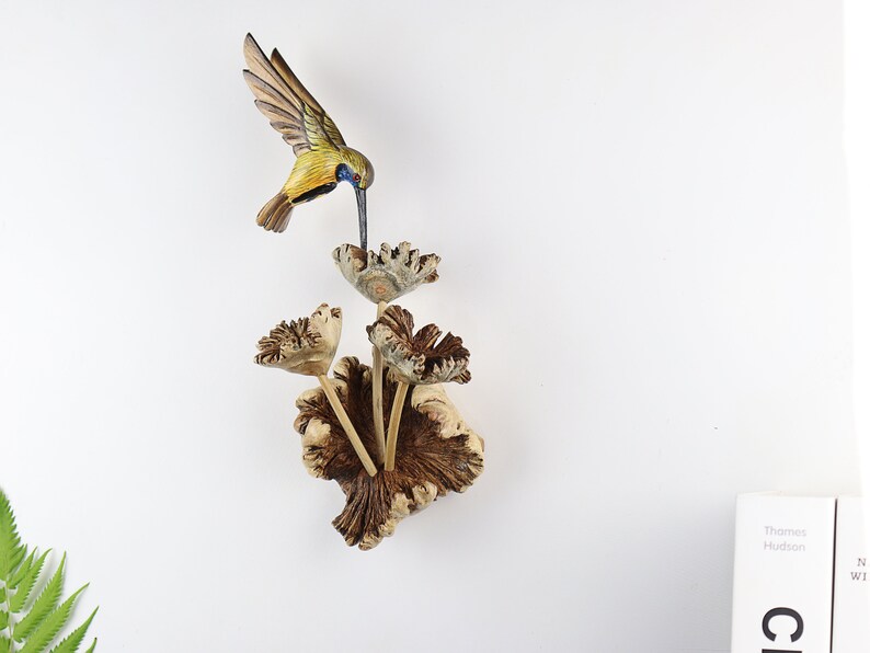 K&ouml;nnte beinhalten: Eine Holzskulptur eines Kolibris in Gelb, Blau und Braun, der &uuml;ber einem Strau&szlig; geschnitzter Holzblumen schwebt. Die Skulptur ist auf einem hellen Holzsockel montiert und schafft ein von der Natur inspiriertes Dekorationsobjekt.