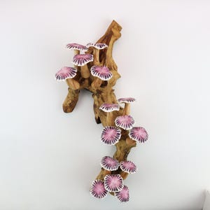 May include: A decorative wooden sculpture featuring a natural, textured branch adorned with several ceramic mushrooms. The mushrooms have white stems and caps with a pink and purple radial pattern. The sculpture is mounted on a white wall.
