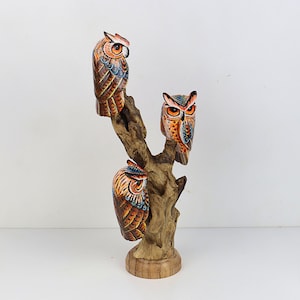 Puede incluir: Figurita de b&uacute;ho de madera tallada a mano con tres b&uacute;hos posados en una rama. Los b&uacute;hos est&aacute;n pintados en tonos marrones, naranjas y azules con detalles intrincados.