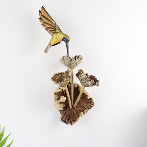 K&ouml;nnte beinhalten: Holzvogel-Skulptur mit ausgebreiteten Fl&uuml;geln, bemalt in Gelb-, Braun- und Blaut&ouml;nen, schwebt &uuml;ber einer Gruppe stilisierter Holzblumen. Die Blumen sind an einer strukturierten, braunen Holzbasis befestigt und schaffen ein von der Natur inspiriertes Wandkunstwerk.