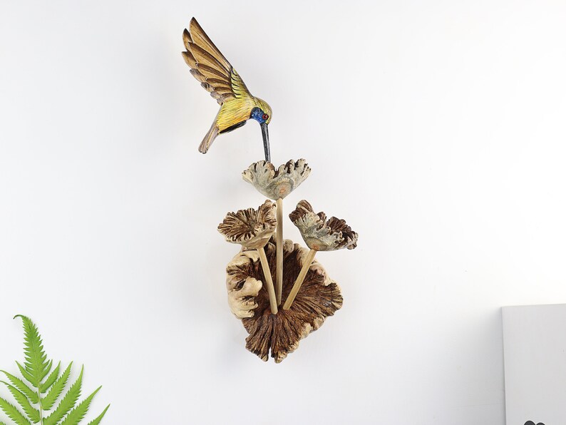 K&ouml;nnte beinhalten: Holzvogel-Skulptur mit ausgebreiteten Fl&uuml;geln, bemalt in Gelb-, Braun- und Blaut&ouml;nen, schwebt &uuml;ber einer Gruppe stilisierter Holzblumen. Die Blumen sind an einer strukturierten, braunen Holzbasis befestigt und schaffen ein von der Natur inspiriertes Wandkunstwerk.
