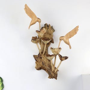 May include: Wooden wall art featuring two carved hummingbirds with outstretched wings, feeding from stylized flowers. The birds and flowers are light brown, mounted on a textured, dark brown wooden base. A nature-themed decorative piece.