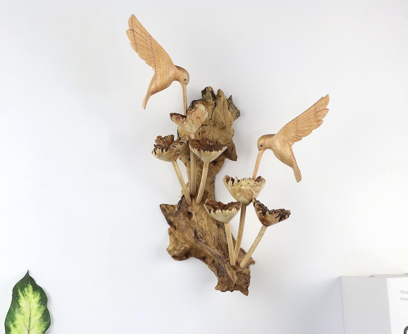 May include: Wooden wall art featuring two carved hummingbirds with outstretched wings, feeding from stylized flowers. The birds and flowers are light brown, mounted on a textured, dark brown wooden base. A nature-themed decorative piece.