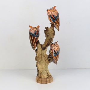 Puede incluir: Tres b&uacute;hos de madera pintados en naranja, marr&oacute;n y azul posados ​​en una rama de madera. Los b&uacute;hos est&aacute;n de espaldas al espectador. La rama est&aacute; unida a una base de madera.