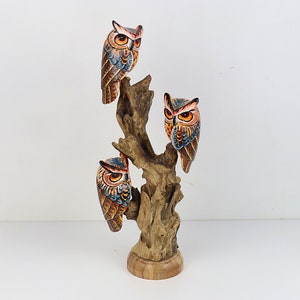 Puede incluir: Conjunto de tres figuras de b&uacute;ho de madera pintadas a mano, posadas en una rama de &aacute;rbol. Los b&uacute;hos est&aacute;n pintados en tonos de marr&oacute;n, naranja y azul con ojos negros. La rama del &aacute;rbol es de color madera natural.