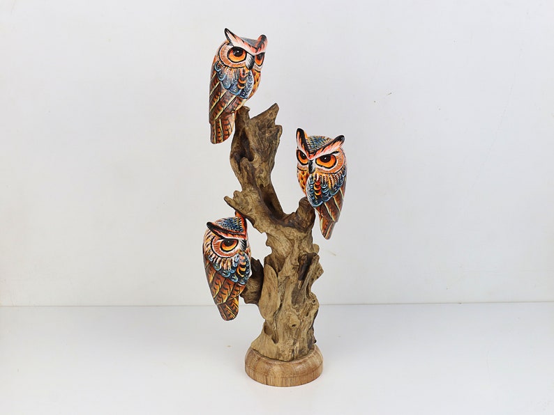 Puede incluir: Conjunto de tres figuras de b&uacute;ho de madera pintadas a mano, posadas en una rama de &aacute;rbol. Los b&uacute;hos est&aacute;n pintados en tonos de marr&oacute;n, naranja y azul con ojos negros. La rama del &aacute;rbol es de color madera natural.