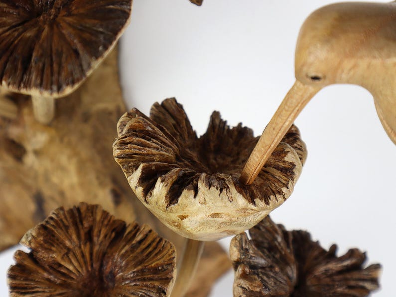 May include: A wooden sculpture featuring a bird with a long beak, reaching into a carved wooden mushroom. The mushrooms are brown with textured caps, and the bird is a light brown color. The sculpture is set against a white background.