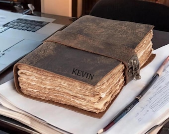 Può includere: Un diario rustico in pelle marrone con il nome "KEVIN" impresso sulla copertina. Il diario è assicurato con un cinturino in pelle e una chiusura in metallo. Una matita e dei fogli sono accanto al diario, suggerendo un tema di scrittura o schizzi.