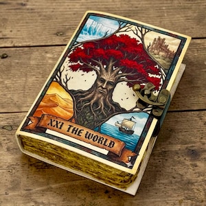Puede incluir: Un diario hecho a mano con una ilustración detallada en la portada que presenta un árbol con rostro, una escena desértica, un barco y un castillo. La portada dice "XXI THE WORLD" en una pancarta. El diario tiene un cierre de metal y páginas envejecidas.