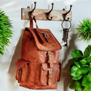Puede incluir: Una mochila de cuero marrón con múltiples bolsillos y hebillas. La mochila está colgada en un perchero de madera con un juego de llaves colgando de un gancho.