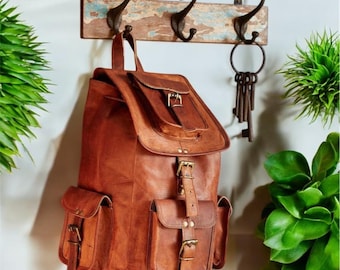 May include: A brown leather backpack with a top flap, a central buckle strap, and two side pockets with buckle closures. The backpack hangs from a rustic wooden wall rack featuring three dark metal hooks and a set of hanging metal keys.