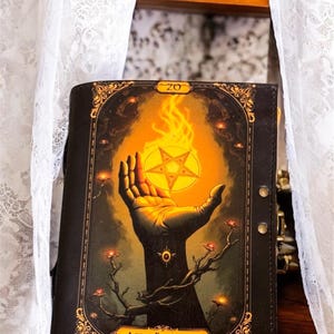 Puede incluir: Un diario encuadernado en cuero negro con una cubierta adornada con detalles dorados que presenta una ilustración de una mano sosteniendo un pentagrama con llamas. El diario tiene el texto "Ace of Pentacles" y "20" en la portada.