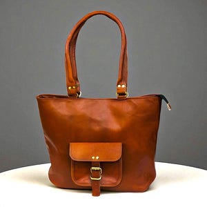 May include: A brown leather tote bag with a front pocket and a buckle fastening. The bag has two top handles and a zip closure. The bag is on a white surface.