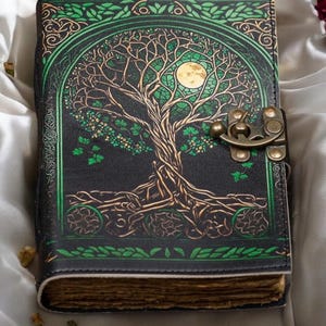 Puede incluir: Un diario de cuero negro con un diseño de árbol de la vida verde y dorado. El diario tiene un cierre de broche y una luna en el árbol.