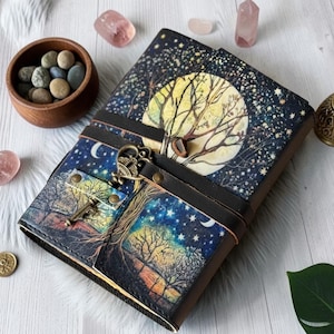 May include: A rectangular journal with a dark cover featuring a night sky, a large yellow moon, and bare trees. A dark brown leather strap wraps around the center, secured by a metallic antique-style key ornament with brass-colored rivets.
