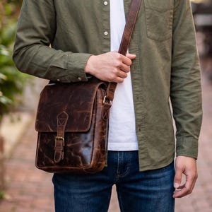 May include: A dark brown leather messenger bag with a vertical flap, a single buckle closure, and an adjustable shoulder strap. The bag features a distressed finish and is worn over an olive green button-down shirt and a white t-shirt with blue jeans.