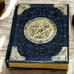 Könnte beinhalten: Ein dunkelblaues Buch mit einem goldenen Pentagramm-Emblem und Eckakzenten. Das Buch hat geprägte Blumenmuster und goldfarbene Kanten. Es liegt auf einer Holzoberfläche, mit anderen dekorativen Gegenständen.
