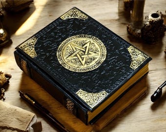 May include: A black book with gold accents and a gold pentagram emblem. The book has ornate corner designs and gold-edged pages. The book is resting on a wooden surface, with other objects in the background.