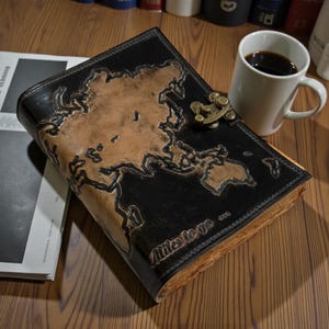 May include: A leather journal with a world map design. The journal is black with a brown map and a brass clasp. The text "Miles to go" is embossed on the cover. A white mug of coffee sits nearby.