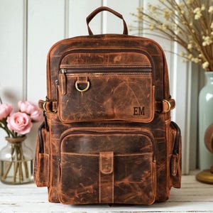 May include: A brown leather backpack with a distressed finish, featuring multiple zippered compartments, a top handle, side pockets, brass-toned hardware, and the initials EM embossed on the front pocket. The design includes a flap closure.