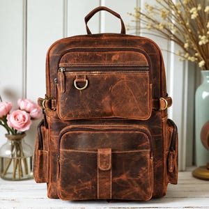 May include: A brown leather backpack featuring a top handle, two main zippered compartments, and two side pockets. The front includes a zippered pocket with a brass D-ring and a lower pocket with a flap closure. The leather has a distressed finish.